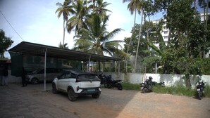 A parking area with a white car and several motorcycles, surrounded by palm trees and a building with a covered structure. - Hotel Poptavern Townbridge (Thiruvananthapuram)