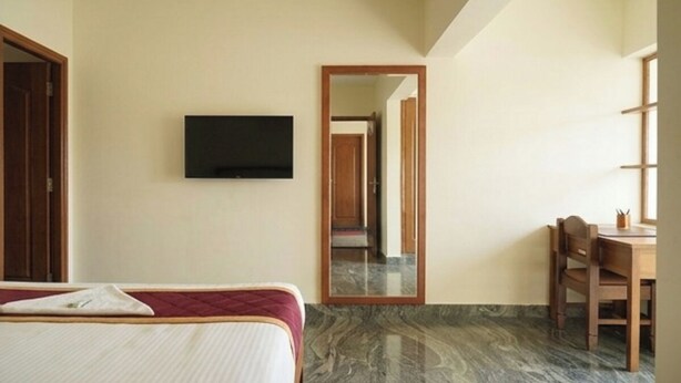 A hotel room with a bed, a television mounted on the wall, a mirror, a wooden chair, and a table. - Hotel Poptavern Townbridge (Thiruvananthapuram)
