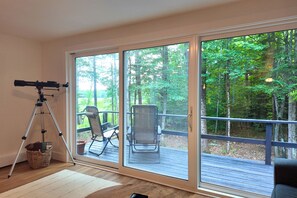 A room with a telescope on a tripod, a wicker basket, and a chair on a deck with a view of a forest.