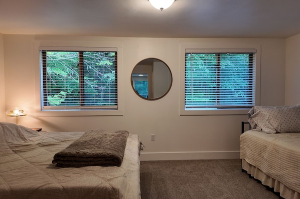 A bedroom with two beds, a round mirror, and windows with blinds.
