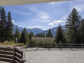 Ein gepflasterter Bereich mit Holzzaun, einer Bank und Blick auf Berge und Bäume.