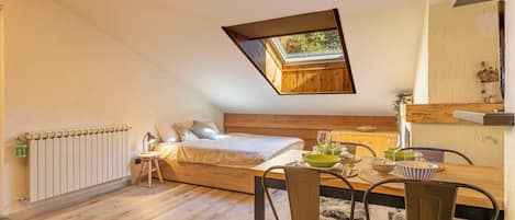 Un dormitorio acogedor con techo inclinado y tragaluz, una cama de madera, un taburete pequeño y una mesa de comedor con sillas.