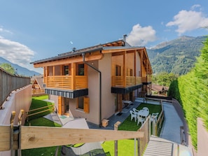 A two-story wooden chalet with a balcony, a small patio with outdoor furniture, and a view of a mountainous landscape.