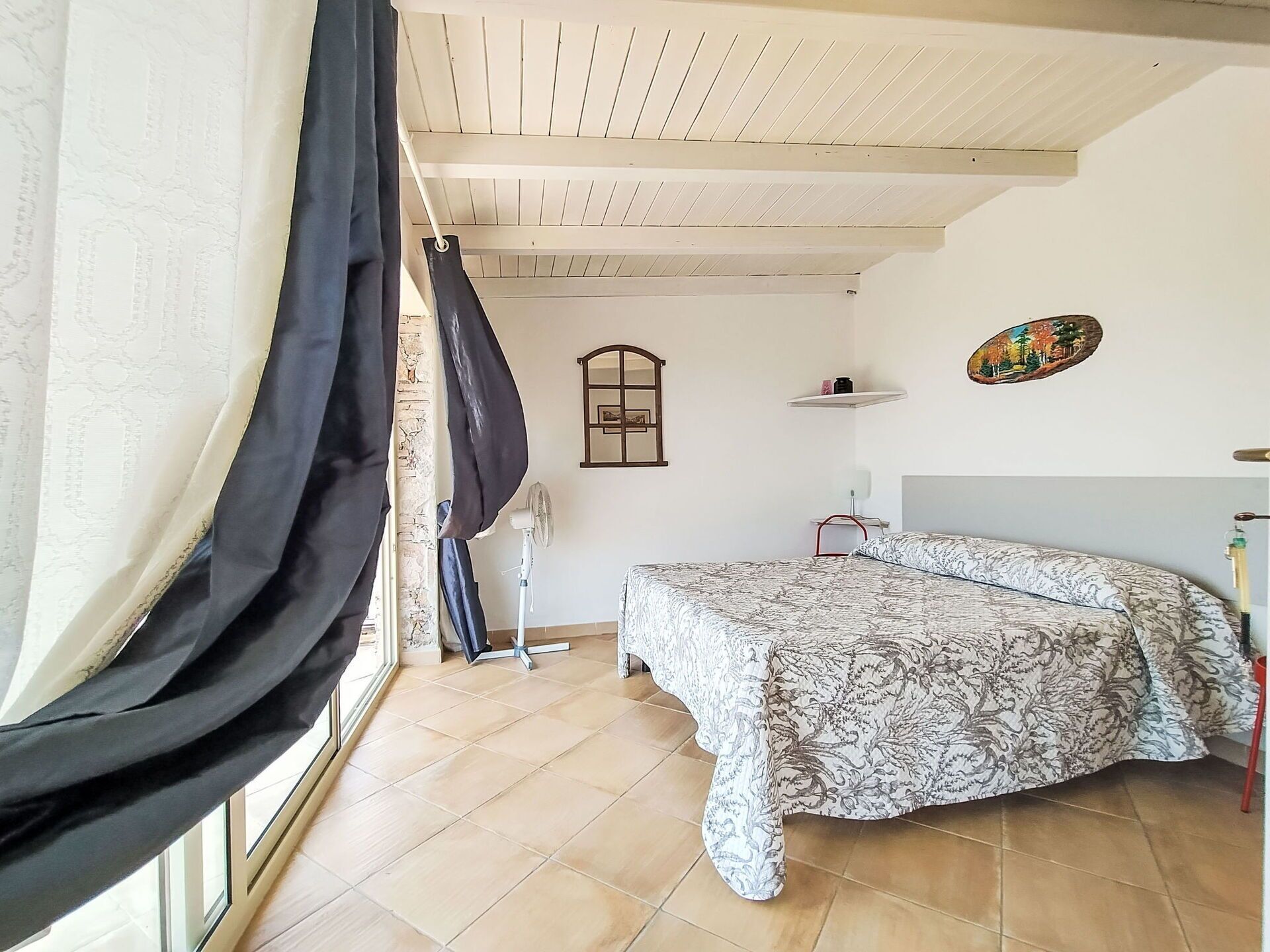 A bedroom with a bed, a window with curtains, a fan, a shelf with items, and a wall decoration.