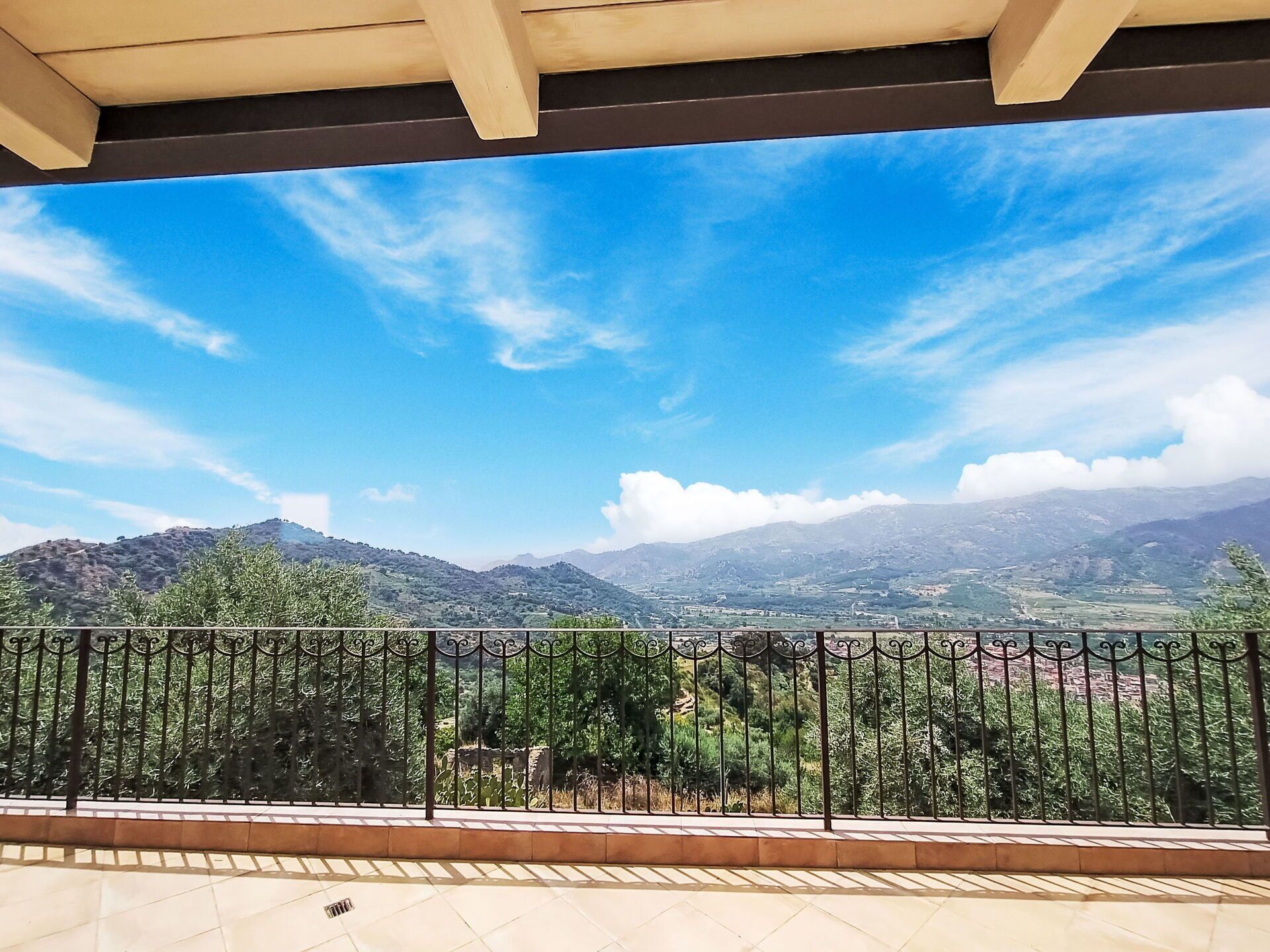 Un balcone con ringhiera metallica, pavimento piastrellato e vista su un paesaggio montuoso sotto un cielo sereno.