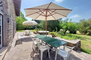 Eine Terrasse im Freien mit einem grünen Tisch, weißen Stühlen, einem großen, beigen Sonnenschirm und einer Steinmauer.