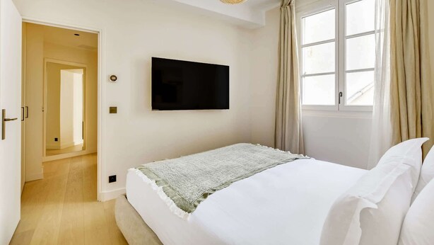 Una camera da letto con un letto, una televisione a parete, una finestra con tende e un'uscita verso un'altra stanza.