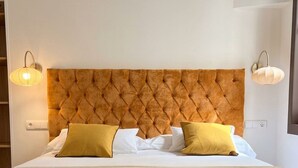 Habitación de hotel con tablero de cabecera capitonado, dos lámparas de noche, cama con ropa blanca y dos almohadas, y un banco al pie de la cama.