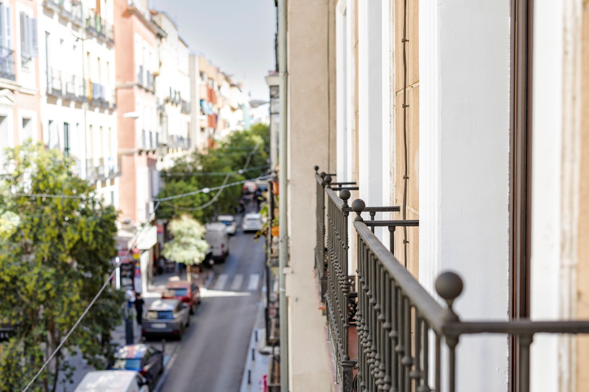 Vista da rua a partir de uma varanda com grade de metal, edifícios, carros estacionados e árvores.