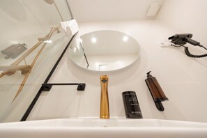 Un baño con un lavamanos blanco, un espejo redondo, un cabezal de ducha manual, un secador de pelo y una botella de loción.