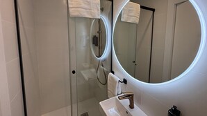Un baño moderno con ducha de vidrio, un espejo redondo con luz integrada y un lavamanos blanco con un solo grifo.