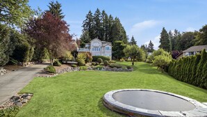 Ein gepflegter Rasen mit Trampolin, umgeben von verschiedenen Bäumen und Sträuchern, und ein Haus im Hintergrund.