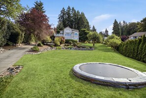 Ein gepflegter Rasen mit Trampolin, umgeben von verschiedenen Bäumen und Sträuchern, und ein Haus im Hintergrund.