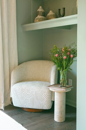 A corner room with a patterned armchair, a small table with a vase of flowers, and a shelf with decorative items. - Livin' La Vida Lakehouse (Henrico)