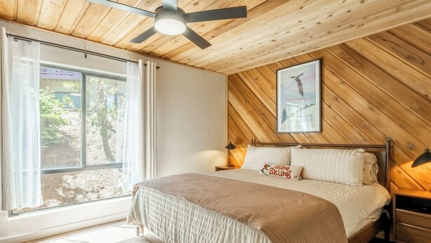 Ein Schlafzimmer mit einem Bett, Deckenventilator, Nachttisch, Lampe, einem Fenster mit Vorhängen und einem gerahmten Bild an der Wand.