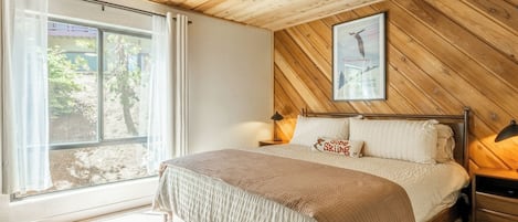 Un dormitorio con cama, ventilador de techo, mesita de noche, lámpara, ventana con cortinas e imagen enmarcada en la pared.