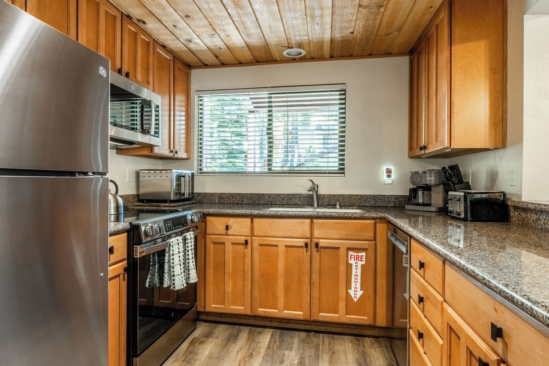 A kitchen with wooden cabinets, stainless steel appliances, a window with blinds, and a fire extinguisher sign.