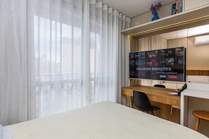 A hotel room with a large bed, a desk with a computer, a TV displaying a movie poster, and sheer curtains. - Modern Studio with a View in Downtown Curitiba - Luxury (Curitiba)