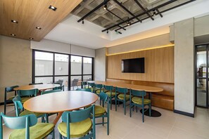 Un intérieur moderne avec des tables et des chaises rondes, un téléviseur à écran plat fixé sur un mur en panneaux de bois, et de grandes fenêtres laissant entrer la lumière naturelle.