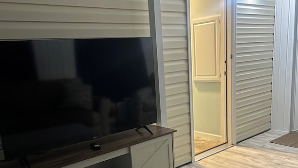A living room with a flat-screen TV on a wooden stand, a grey sofa, and a sliding glass door leading to a garage. - Cozy 1-bedroom tiny home in resort setting (Citra)