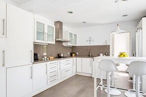 A modern kitchen with white cabinetry, a built-in stove, and a breakfast bar with bar stools. - VILLA MAGDALENA in Cala Blanca (Cala Blanca)
