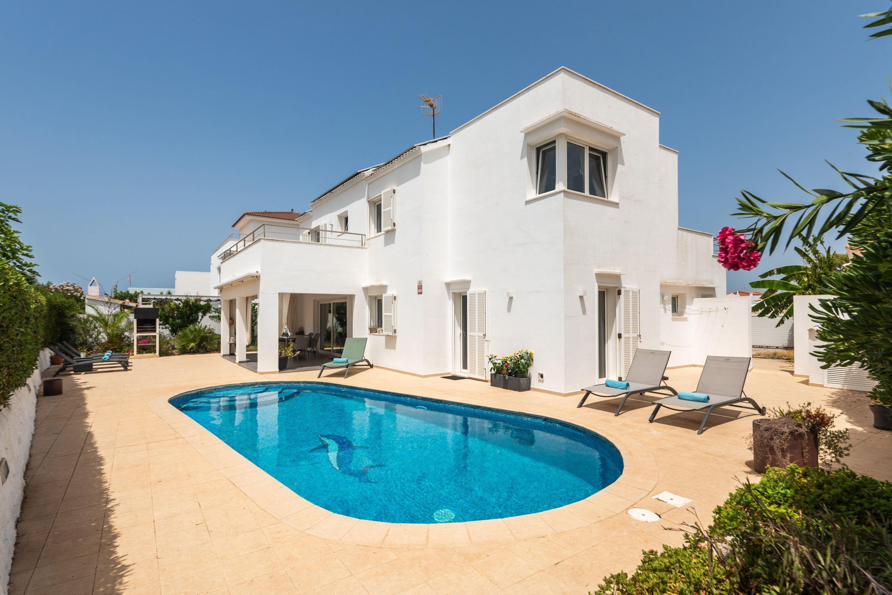 A two-story white building with a swimming pool, outdoor seating, and a clear sky.