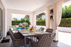 A dining area with a table set for six, surrounded by wicker chairs, with a view of a pool and greenery outside. - VILLA MAGDALENA in Cala Blanca (Cala Blanca)