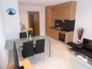 Eine moderne Küche mit Holzausstattung, ein Essbereich mit Glastisch und Stühlen sowie ein Wohnbereich mit Sofa und Glastisch.