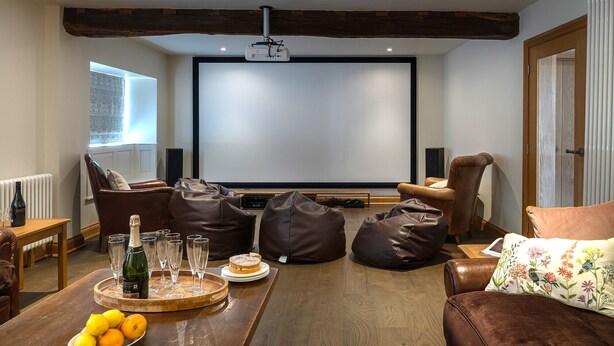 A cozy living room with a wooden coffee table, leather bean bags, a brown leather sofa, and a large screen for viewing. - Finest Retreats | Serenity (Painswick)