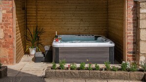 A hot tub in a wooden cabin with a brick wall, potted plants, and a stone pathway. - Finest Retreats | Serenity (Painswick)