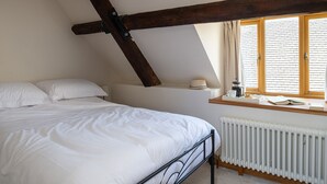 A bedroom with a sloped ceiling, a bed with white bedding, a window with curtains, a radiator, and a shelf with a hat and a mug. - Finest Retreats | Serenity (Painswick)