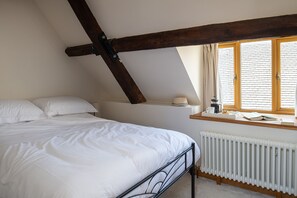 Ein Schlafzimmer mit geneigter Decke, einem Bett mit weißer Bettwäsche, einem Fenster mit Vorhängen, einem Heizkörper und einem Regal mit Hut und Tasse.