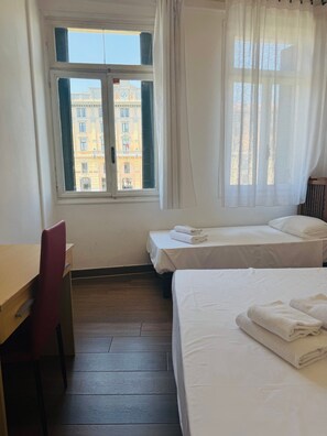 Een hotelkamer met een houten bureau, twee bedden, een stoel en een raam met uitzicht op gebouwen.