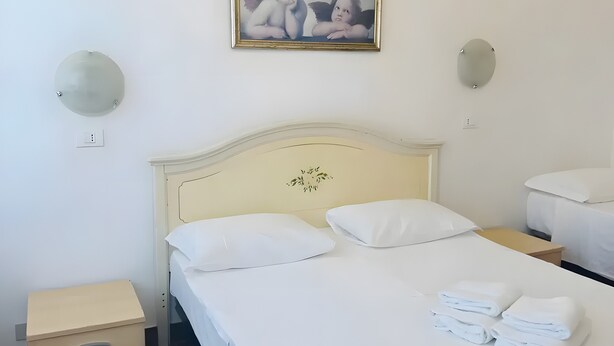 Un letto rifatto con lenzuola e cuscini bianchi, un comodino con una lampada e un quadro appeso alla parete.