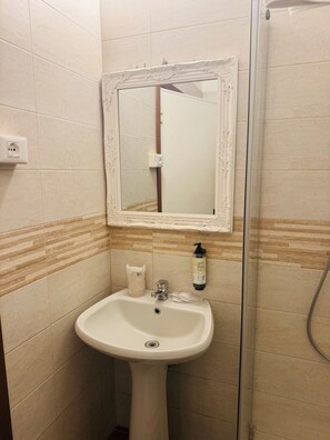 Un baño con lavamanos pedestal, un espejo con marco ornamentado, dispensador de jabón y zona de ducha.