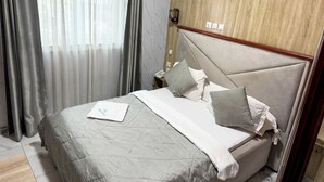 A neatly made bed with a grey bedspread and pillows, a bedside table with a lamp, and a window with curtains.