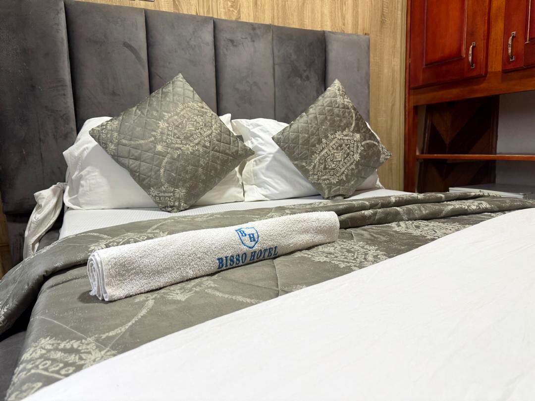 A neatly made bed with a grey and white duvet, two patterned pillows, and a rolled white towel with a logo.