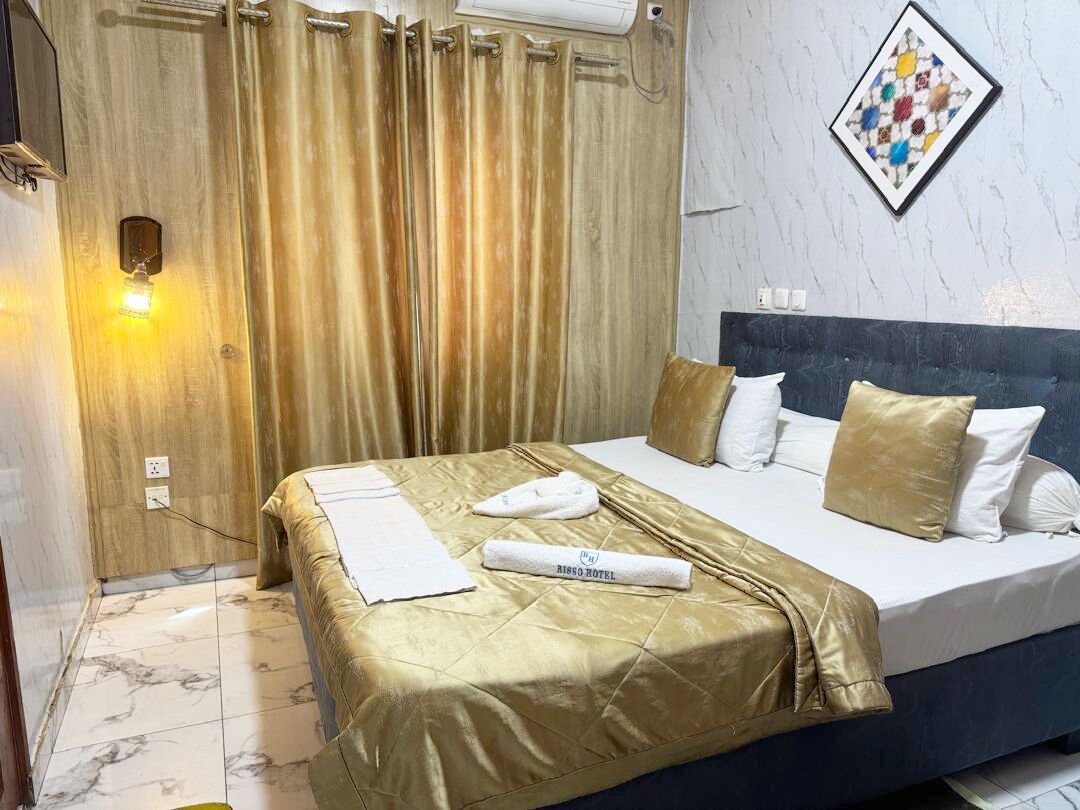 Een hotelkamer met een bed, hoofdeinde, een wandlamp, gordijn en een lijst met een afbeelding.