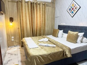 Habitación de hotel con cama, cabecera, luz de pared, cortina e imagen enmarcada.