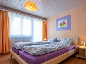 Ein Schlafzimmer mit zwei Betten, einem Fenster mit durchsichtigen Vorhängen, einem Bild an der Wand und einer Nachttischlampe.