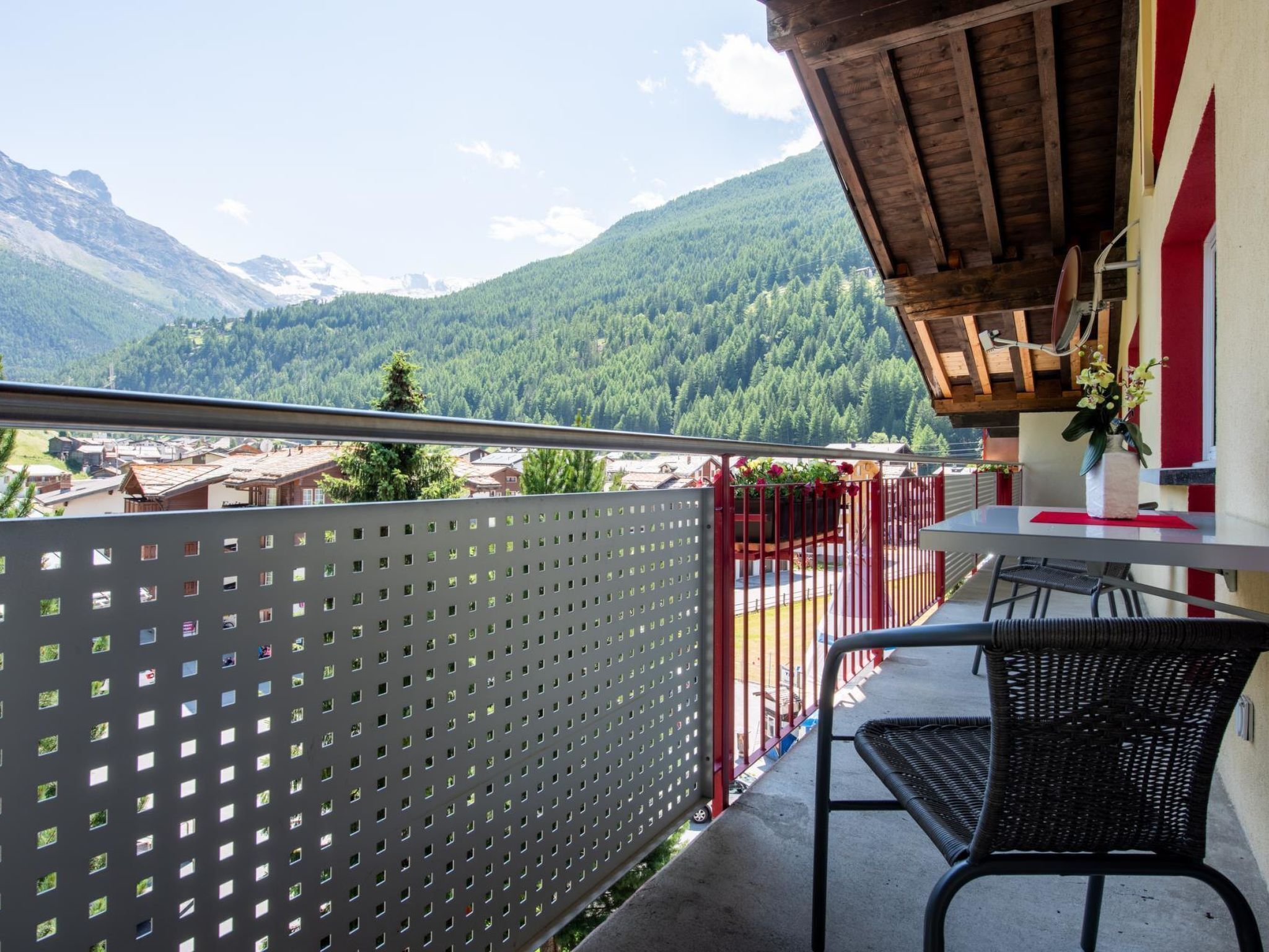 A balcony with a view of a mountain landscape, a table with chairs, and a red railing.
