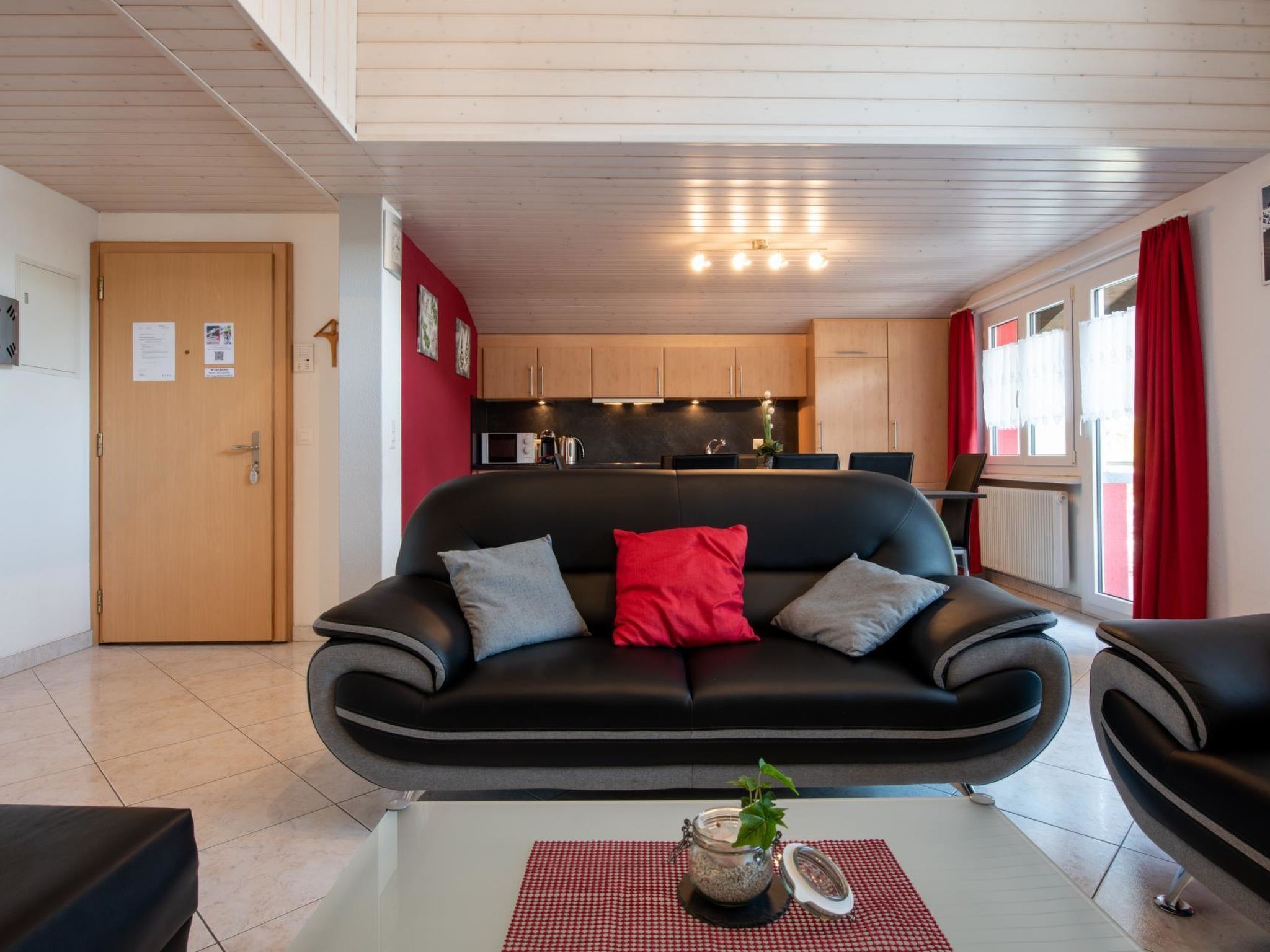 Un salon moderne avec un canapé en cuir noir, une table basse en verre ornée d’une plante et un chemin de table à carreaux rouges et blancs.