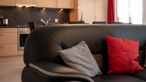 Ett modernt vardagsrum med en svart soffa, en grå och en röd kudde, och ett kök i bakgrunden.