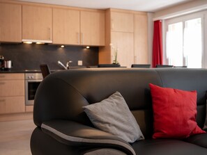 Ein modernes Wohnzimmer mit einer schwarzen Couch, einem grauen und einem roten Kissen, und einer Küche im Hintergrund.
