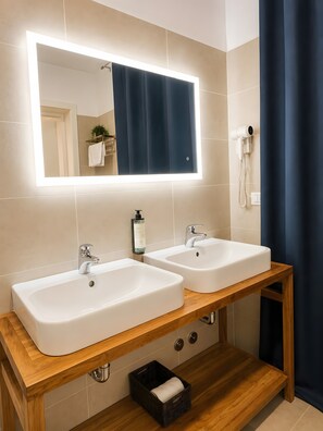 Ein Badezimmer mit zwei weißen Waschbecken, einem Holzwaschtisch, einem Spiegel mit integrierter Beleuchtung, einem Haartrockner und einem dunklen Vorhang.