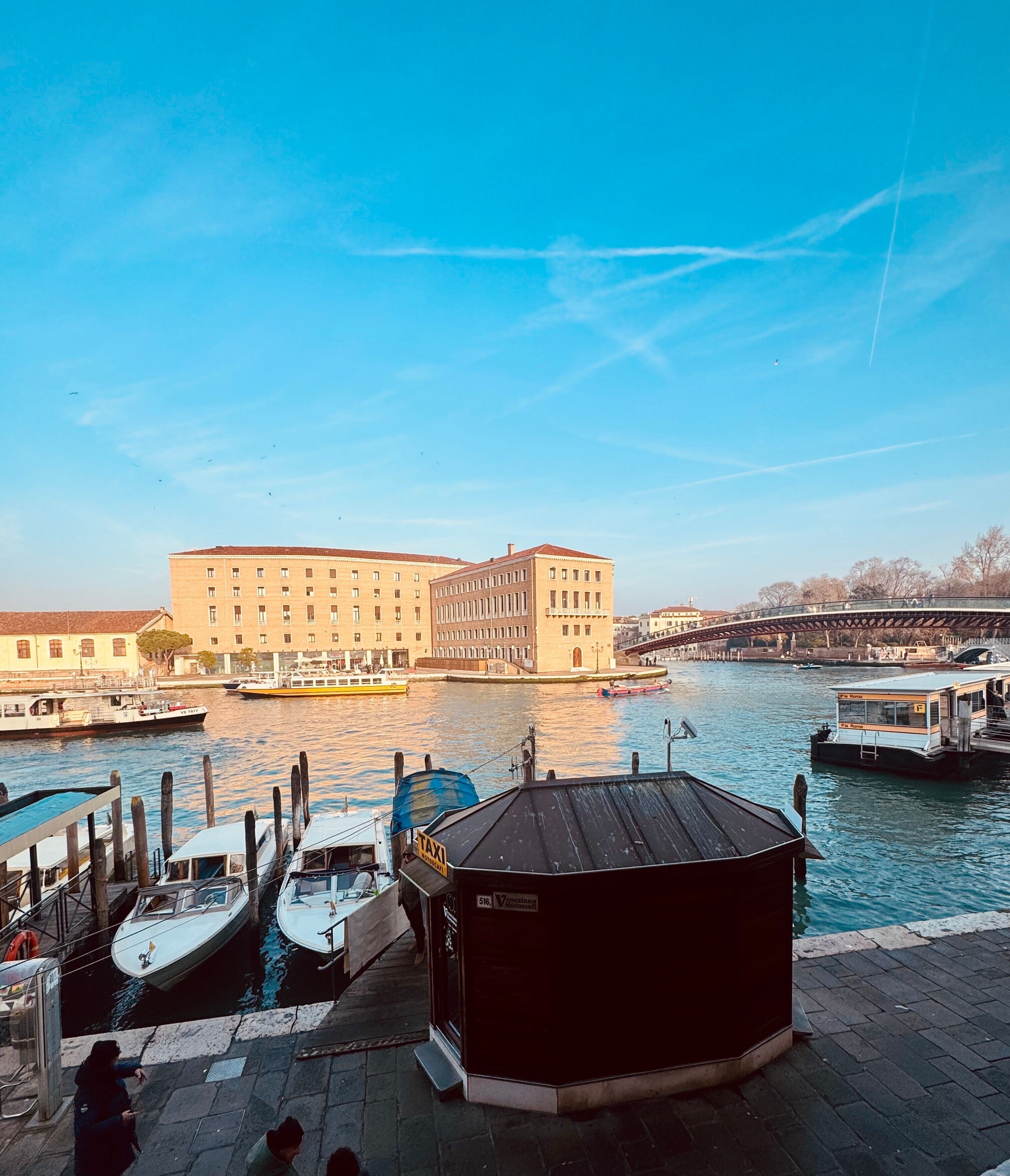 A waterfront view with boats docked, a large building, and a bridge in the background.