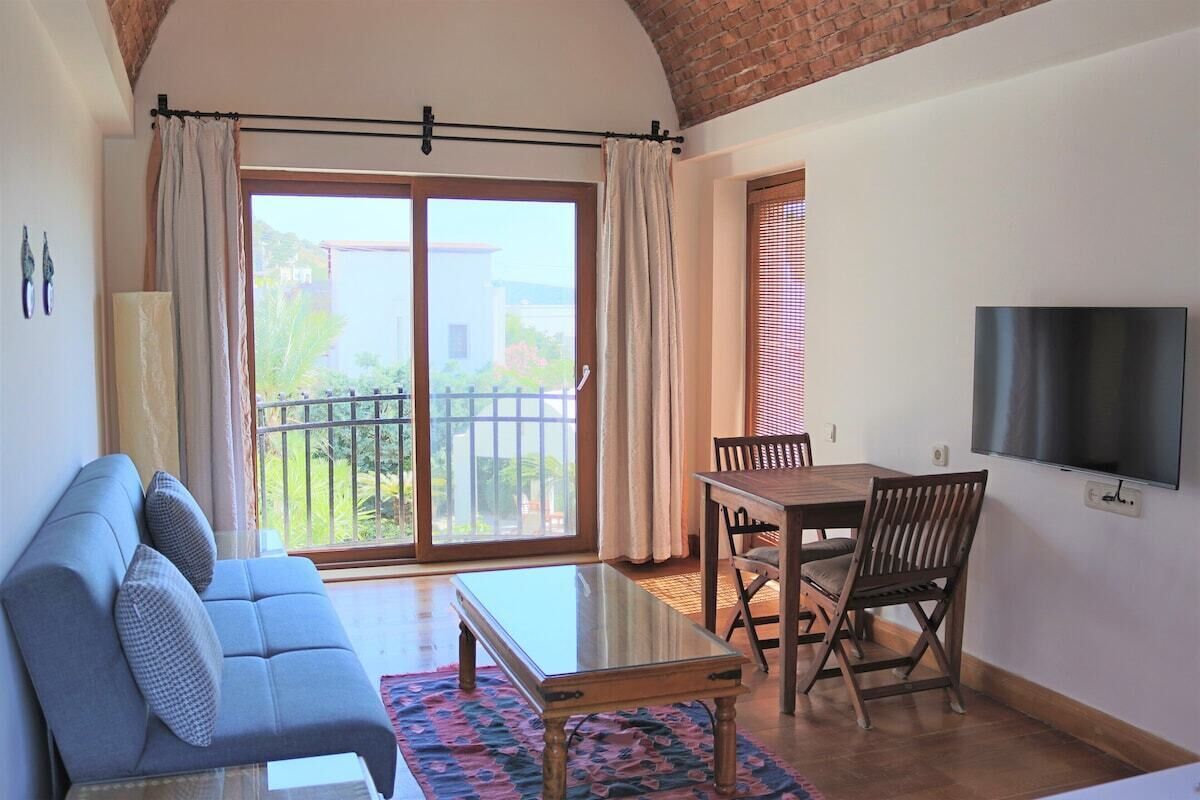 Una sala de estar con sofá, una mesa de centro de vidrio, una mesa de comedor con sillas, un televisor montado en la pared y un ventanal corredero que da a un área exterior.