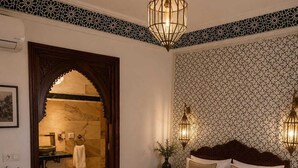 Habitación de hotel con una cama grande, una mesita de noche, una lámpara de pared y un baño visible a través de un arco.
