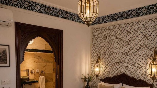 A hotel room with a large bed, a nightstand, a wall-mounted lamp, and a bathroom visible through an archway. - riad el barodienne  (Marrakech)