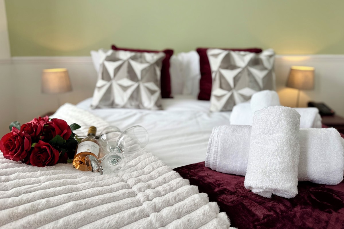 A neatly made bed with white linens, a red rose bouquet, and a bottle of perfume.